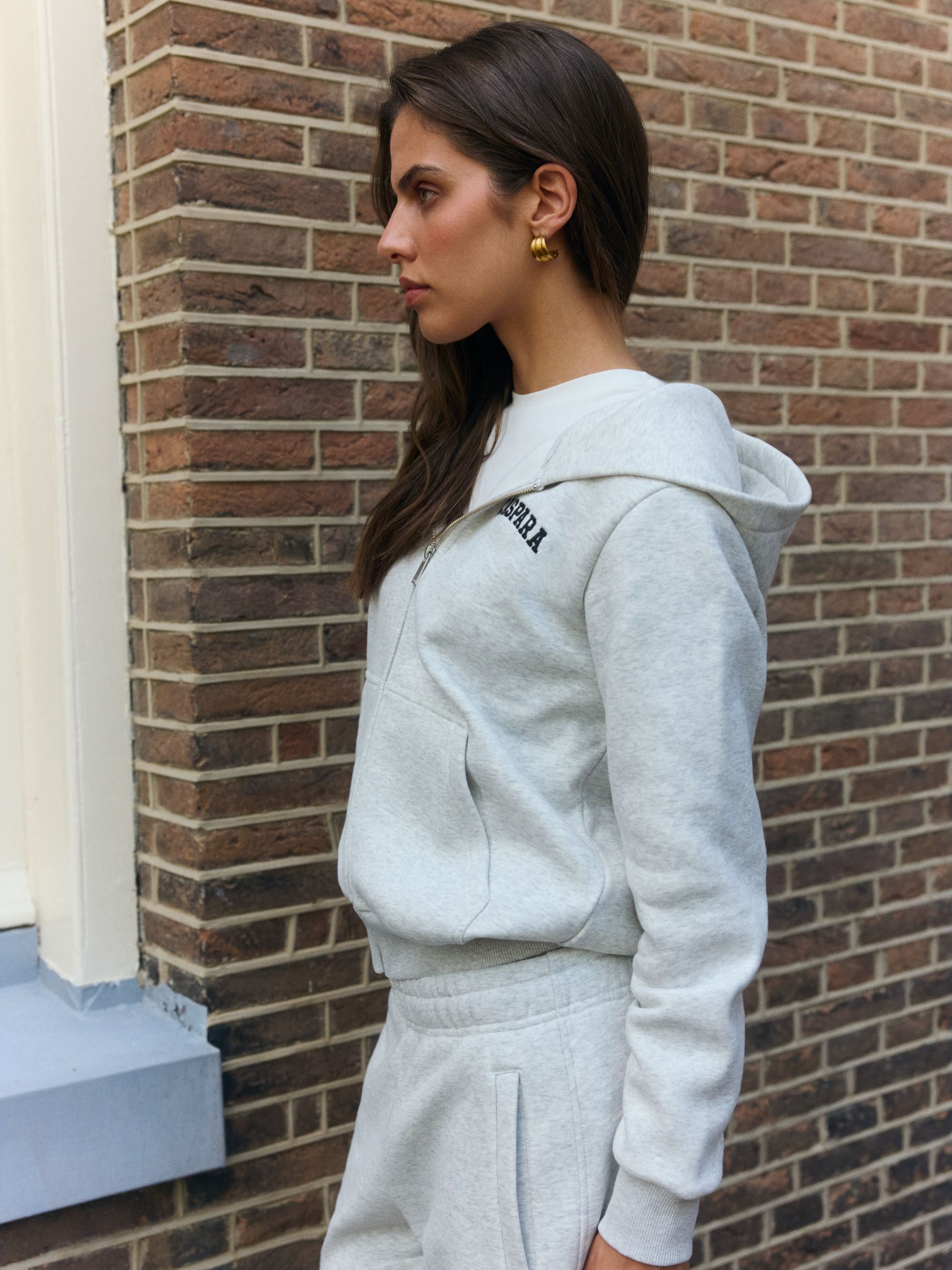 Woman wearing a grey melange CASPARA zip hoodie standing against a brick wall.